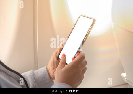 Frau im Flugzeug halten ihr Smartphone, während sie im Flugzeug sitzt. Frau, die im Flugzeug fliegt, benutzt ein Mobiltelefon mit einem weißen Bildschirm. Draht Stockfoto