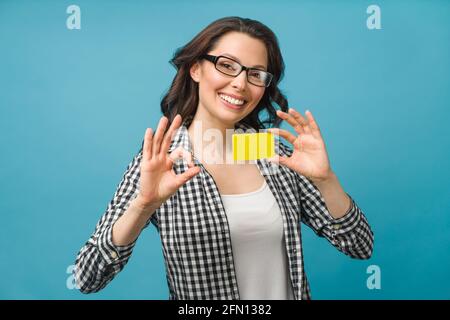 Niedliche lächelnde Frau in Hemd und Brille, zeigt Kreditkarte in der Hand für Finanz- und bargeldlose Gesellschaft Konzept, posiert auf blauem Hintergrund Stockfoto