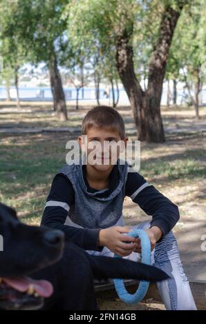 10-jähriger Junge und 11-jähriger labrador für einen Spaziergang. Ein Kind mit einem Haustierspielzeug sitzt und schaut auf die Kamera. Verschwommener schwarzer Hund im Vordergrund. Stockfoto