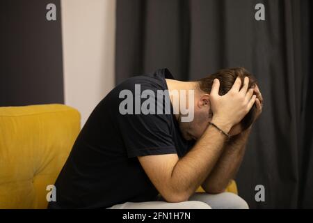 Der unrasierte Kerl sitzt auf dem Sofa und packte seinen Kopf in die Hände An Depressionen leiden Stockfoto