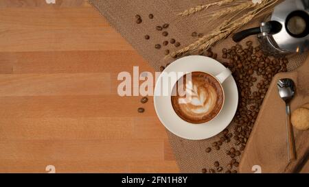 Blick von oben auf den Frühstückstisch mit Latte-Kaffeetasse, Kaffeekocher auf Tischset, dekoriert mit Kaffeebohnen Stockfoto