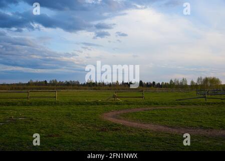 Ein grünes Feld mit einem Holzzaun für Pferde vor dem Hintergrund eines Sonnenuntergangs wolkigen Himmels. Frühlingslandschaft. Stockfoto