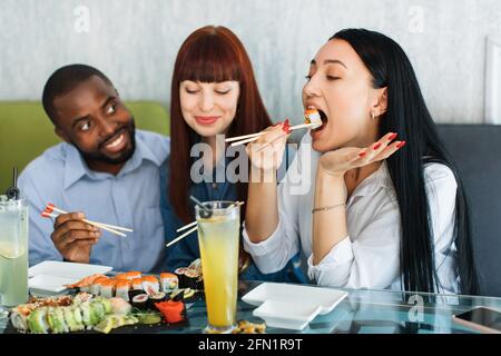 Junge, multiethnische Menschen essen Sushi-Brötchen im asiatischen Restaurant. Glückliche junge Freunde, die ihre Freizeit und Sushi im Café genießen. Konzentrieren Sie sich auf asiatische Brünette Mädchen essen Rolle und mit Holzstäbchen Stockfoto