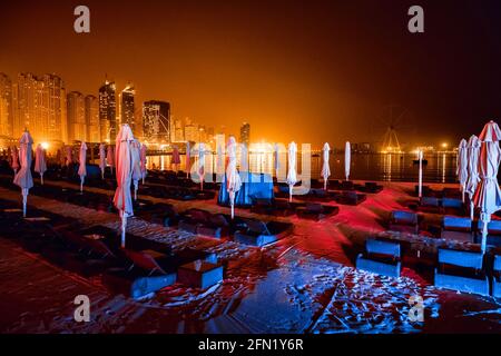 Blick vom Strand in Dubai, mit Liegestühlen und geschlossenen Sonnenschirmen, auf die Wolkenkratzer und das Rad der Unbeweglichkeit Stockfoto