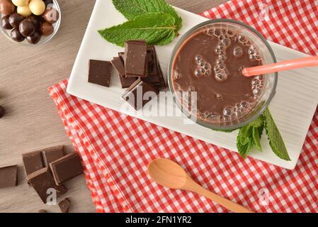 Glas mit Milchschokolade-Shake auf Teller mit Kugeln und Schokoladenportionen auf rot-weiß karierter Tischdecke auf Holzbank. Draufsicht. Stockfoto