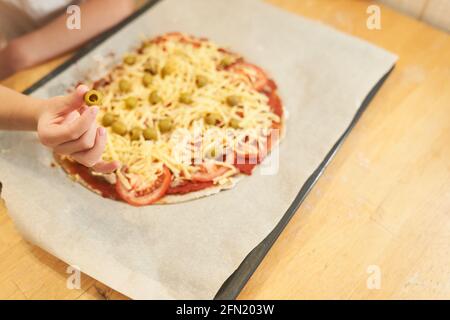 Eine Kinderhand mit einer Olive auf dem Hintergrund einer gesunden glutenfreien hausgemachten Pizza, die zum Backen zubereitet wird. Stockfoto