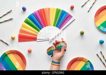 LGBTQ Community PRIDE Monat Regenbogen Konzept flach Lay. Hand mit Band und Sonnenbrillen. Regenbogen-Fan, Party-Pappteller und Trinkhalme. Flach liegend, Draufsicht Stockfoto