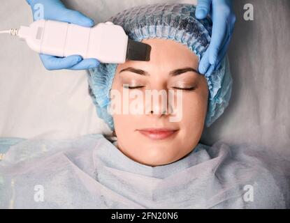 Nahaufnahme der Hände der Kosmetikerin in sterilen Handschuhen mit Ultraschallschrubber während der Reinigung des weiblichen Gesichts der Kundin. Junge Frau in Einweg-Duschhut mit Tiefenreinigung Hautpflege Verfahren in Schönheitssalon Stockfoto