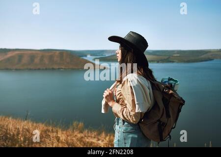 Positive junge Frau im Cowboyhut, die in der Gegend von Bakota durch die grüne Natur spazierengeht. Glückliche Hündin mit Rucksack verbringen Freizeit aktiv. Stockfoto