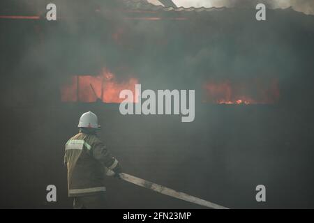 Ein Team von Rettungskräften löscht ein Feuer im Haus. Feuerwehrleute gießen Wasser auf die tobenden Flammen. Rauch und Smog aus dem brennenden Gebäude Stockfoto