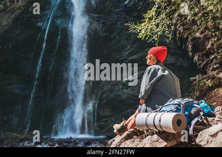 Lächelnde Frau mit Rucksack in aktiver Trekkingkleidung, die in der Nähe des Gebirgswasserfalls sitzt und die spritelnde Kraft der Natur genießt. Reise Stockfoto
