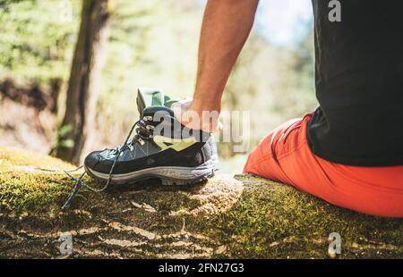 Mann, der auf dem gefallenen Baumstamm über dem Bergwaldbach sitzt, während er seine Trekkingstiefel trocknet. Stiefel Nahaufnahme Foto. Aktive Menschen auf Reisen, Stockfoto