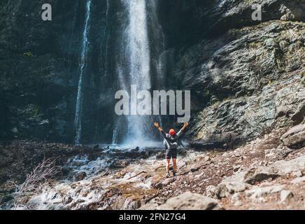 Eine Frau mit rotem Hut und aktivem Trekking Kleidung, die in der Nähe des Gebirgswasserfalls stand, erhob die Arme Und genießen Sie den planschenden Naturpow Stockfoto