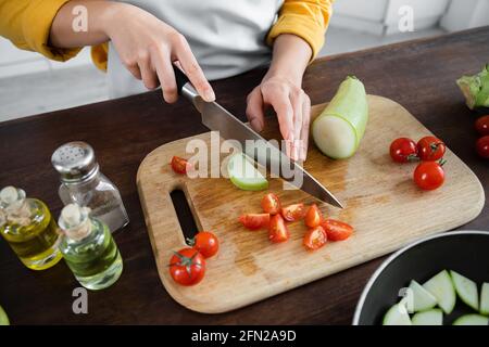 Teilansicht einer Frau, die Zucchini in der Nähe von geschnittenen Kirschtomaten schneidet Auf dem Schneidbrett Stockfoto