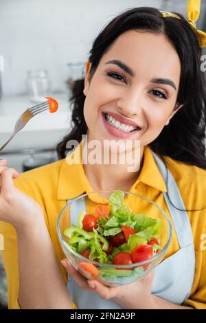 Fröhliche junge Frau, die die Gabel mit Kirschtomate in der Nähe des Salats hält In der Schüssel Stockfoto