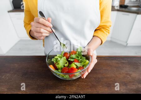 Beschnittene Ansicht einer jungen Frau in der Schürze, die die Gabel in der Nähe hält Salat in einer Schüssel Stockfoto
