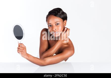 Junge Afrikanerin im Studio, die Körperpflege und Kontrolle macht Ihr Gesicht für Pickel und Flecken mit Spiegel für junge Menschen Und schöne Haut Stockfoto