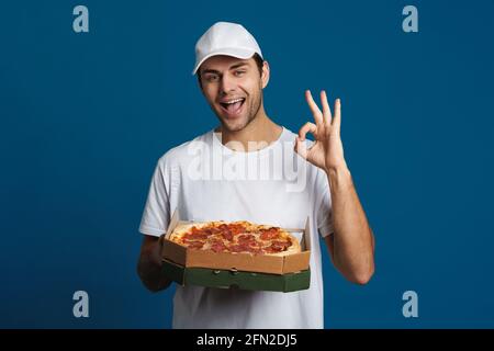 Glücklicher kaukasischer Lieferer, der ein Friedenszeichen zeigt, während er mit ihm posiert Pizza auf blauem Hintergrund isoliert Stockfoto