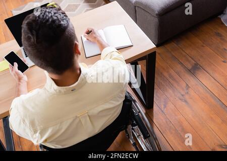 Overhead-Ansicht eines deaktivierten Mannes, der in einem Notebook schreibt, während er die Hand hält Smartphone mit leerem Bildschirm Stockfoto