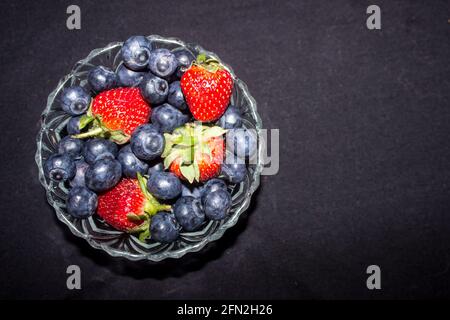 Eine kleine Glasschüssel, gefüllt mit einer Mischung aus Heidelbeeren und Erdbeeren, vor schwarzem Hintergrund Stockfoto