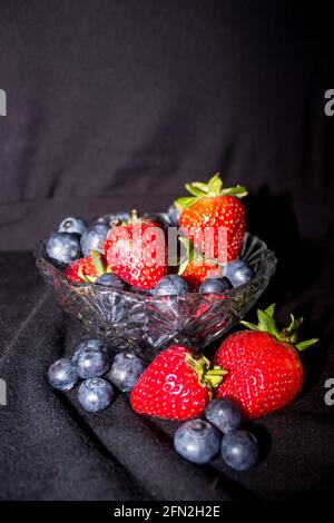 Eine kleine Glasschale gefüllt und umgeben von einer Mischung aus Heidelbeeren und Erdbeeren, vor schwarzem Hintergrund Stockfoto