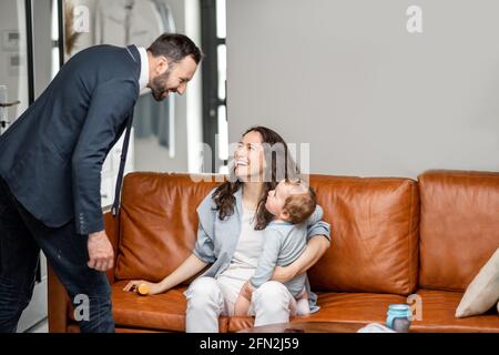 Junge Mutter sitzt mit neugeborenen Sohn auf dem Sofa und kümmern oder spielen mit einem Kind, während Vater und Mann zur Arbeit gehen und sagen Auf Wiedersehen. Stockfoto