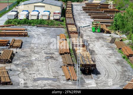 Lange und dicke, rostbedeckte Stahlstäbe, die draußen auf einem Kiesplatz gestapelt sind, sichtbare Eisenbahnschienen und Waggons, von oben betrachtet. Stockfoto