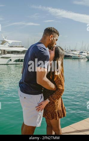 Schwarzer Freund küssen hispanische Freundin auf der Vorderseite beim Umarmen Sie am Hafen mit Booten Stockfoto