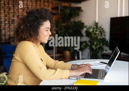 Seitenansicht einer intelligenten und cleveren, positiv-afroamerikanischen Studentin, die einen Laptop für das Fernstudium verwendet. Junge Unternehmerin mit gemischter Rasse, die von zu Hause aus arbeitet, arbeitet gern als freiberufliche Mitarbeiterin Stockfoto