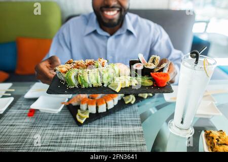 Nahaufnahme eines bunten, vielfältigen Sets von Sushi-Rollen auf einem großen schwarzen Teller in den Händen eines lächelnden, glücklichen jungen Afroamerikaners, der im modernen Stadtrestaurant am Tisch sitzt Stockfoto