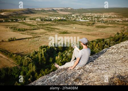 Rückansicht eines jungen Mannes, der Tee oder Kaffee in den Bergen trinkt. Reisender Mann mit Bart hält in den Händen eine Tasse Tee, nach dem Wandern. Reisen, Lifestyle, VA Stockfoto
