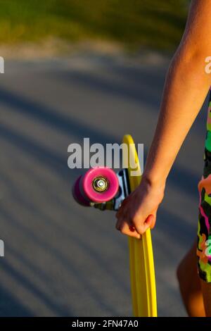 Defokussieren Mädchen mit gelben Penny Board. Rückansicht. Jugend-Hipster-Kultur. Nahaufnahmen von Kinderhänden, die tagsüber ein kurzes Cruiser-Pennyboard im Freien halten. Acti Stockfoto