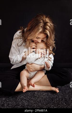 Stilvolle blonde behaarte junge Mutter sanft sitzend und hält Baby Mädchen. Attraktive Frau lächelt und umarmt ihre kleine Tochter im Dunkeln Stockfoto