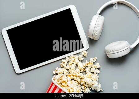 Weißes digitales Tablet mit schwarzem Bildschirm und Kopierbereich mit Popcorn und weiße Kopfhörer auf grauem Hintergrund Stockfoto