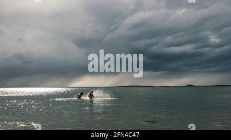 Zwei Freunde spielen im Meer. Langer Schuss von zwei Männern, die Wasser spritzen und in den kalten Gewässern der Nordsee baden. Sommersturm am Horizont. Stockfoto