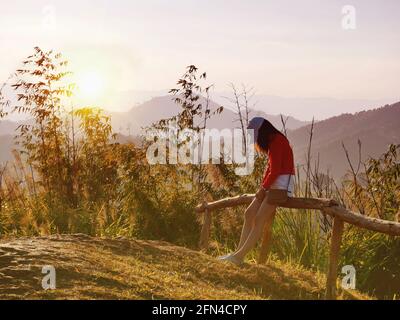 Eine Frau mit rotem Pullover sitzt am Abend in trauriger und einsamer Stimmung auf dem Hügel auf einem Holzzaun. Stockfoto