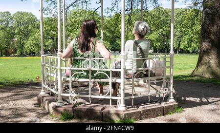 Zwei Frauen verschiedener Generationen sitzen in der Pergola im Park, im Schatten an sonnigen Sommertagen. Blick von hinten. Stockfoto