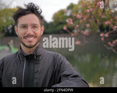 Ein lächelnder Mann mit Brille posiert vor einem japanischen See. Touristen, die Tokio im Frühjahr besuchen. Süßer Mann und blühende Kirschbäume. Stockfoto