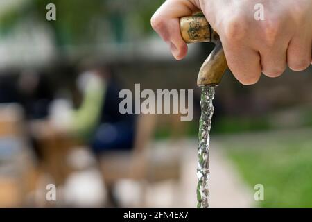 Wasserstrahl, der aus einem Brunnen kommt. An einem heißen Sommertag drückt eine Hand den Wasserhahn eines öffentlichen Brunnens. Wasserabfall und -Verwertung für die Umwelt. Stockfoto