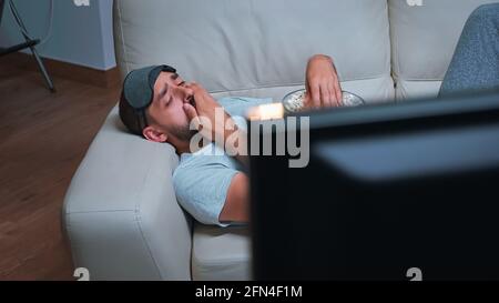 Kaukasischer Mann mit Augenschlafmaske, der Popcorn isst, während er eine Unterhaltungs-tv-Show ansieht. Müder Mann, der während eines Filmfilms spät in der Nacht in der Küche vor dem Fernseher sitzt Stockfoto