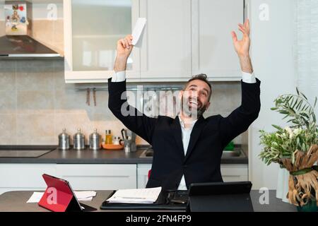 Porträt eines fröhlichen jungen Geschäftsmannes, der beide Arme für einen Deal im Morgengrauen hebt. Ausdrucksstarker Hausangestellter freut sich in der Küche. Erfolgreiches Meeting. Stockfoto