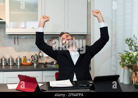 Porträt eines fröhlichen jungen Geschäftsmannes, der beide Arme für einen Deal im Morgengrauen hebt. Ausdrucksstarker Hausangestellter freut sich in der Küche. Erfolgreiches Meeting. Stockfoto