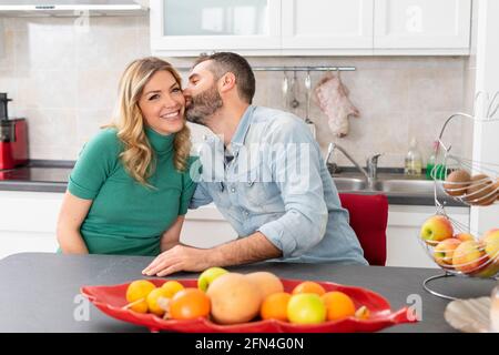Glückliches Paar küsst sich in der Küche. Das Brautpaar sitzt am Küchentisch. Freund küsst seine blonde Freundin. Guten Morgen Kuss. Stockfoto