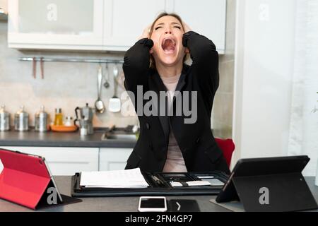 Verzweifelt überarbeitete Geschäftsfrau schreit und schreit in der Küche. Die Frau hält ihren Kopf in den Händen, während sie an einem Nervenzusammenbruch leidet. Stockfoto