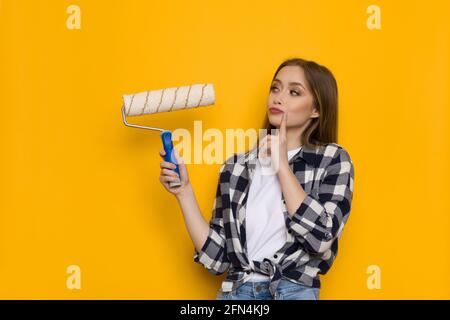 Junge blonde Frau im Holzfällerhemd hält die Rolle der Farbe und denkt nach. Aufnahme auf gelbem Hintergrund Stockfoto