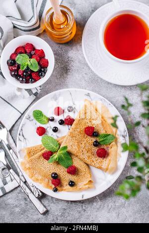 Leckeres Crepes Frühstück auf grauem Beton-Tisch Hintergrund. Orthodoxer Urlaub Maslenitsa. Pfannkuchen mit Beeren schwarze Johannisbeere, Himbeere, Glas Honig Stockfoto
