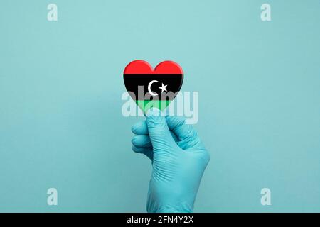 Hände tragen schützende OP-Handschuhe, die das Herz der Libyschen Flagge halten Stockfoto