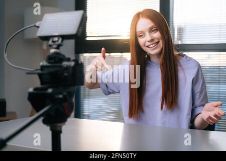 Nahaufnahme einer positiven jungen Bloggerin, die mit einer professionellen Kamera und einer Softbox am Schreibtisch vor dem Fenster über ein neues Telefon spricht. Stockfoto