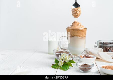 Transparentes Glas mit Kaffee und Milchdessert und einem Löffel mit Creme auf weißem Hintergrund. Dalgon-Kaffee. Horizontale Ansicht, Kopierbereich. Stockfoto