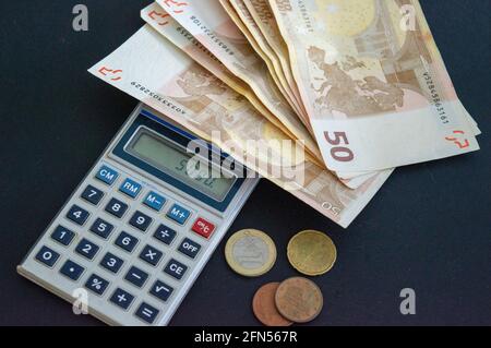 Taschenrechner mit 50-Euro-Banknoten und -Münzen auf grau-schwarzem Hintergrund. Stockfoto
